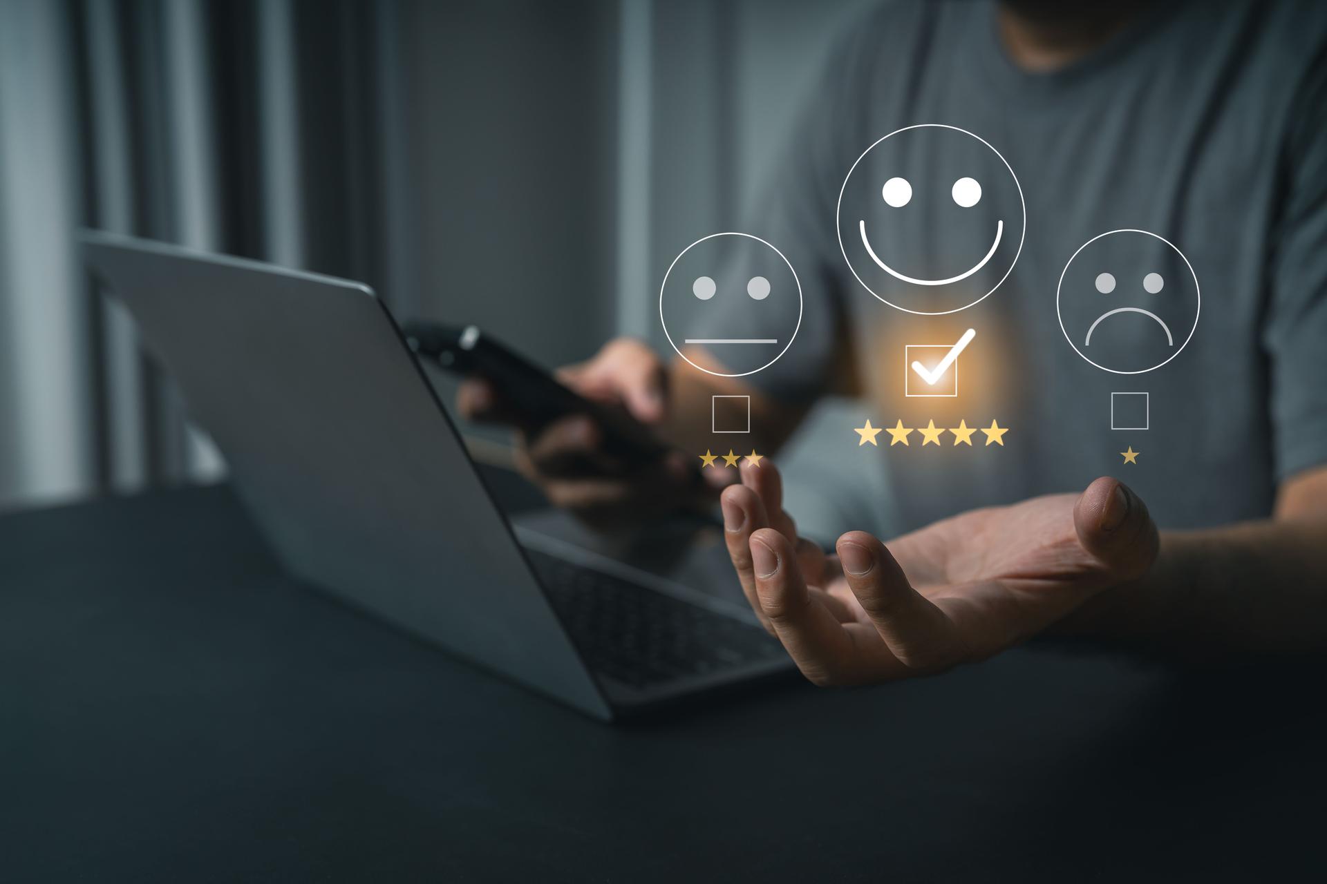 Customer Feedback Concept with Smiley Faces and Rating Stars Over a Person Holding a Phone Near a Laptop in Modern Workspace Environment